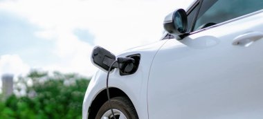 Focus closeup electric vehicle plugged in with EV charger device from blurred background of public charging station powered by renewable clean energy for progressive eco-friendly car concept.