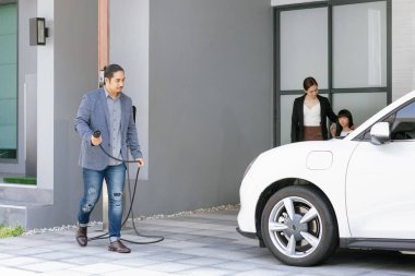 Progressive young parents and daughter with electric vehicle and home charging station. Green and clean energy from electric vehicles for healthy environment. Eco power from renewable source at home.