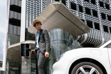 Below view of progressive businessman with electric car recharging at public charging station at modern city residential building. Eco friendly rechargeable EV car powered by alternative clean energy.