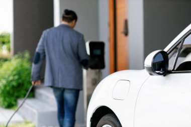 Focus electric car charging at home charging station with blurred progressive man walking in the background. Electric car using renewable clean for eco-friendly concept.