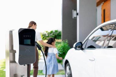 Arka planda yürüyen ilerici kadın ve kız ile elektrikli araç için temiz enerjiyle elektrik santraline odaklan. Elektrikli araba konsepti için ev şarj istasyonu