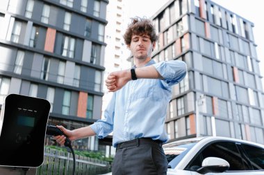 Businessman with smartwatch at modern charging station for electric vehicle with background of residential buildings as concept for progressive lifestyle of using eco-friendly as alternative energy.
