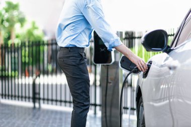 Closeup progressive man holding EV charger plug from public charging station for electric vehicle with background of residential building as concept eco-friendly sustainability energy car concept.