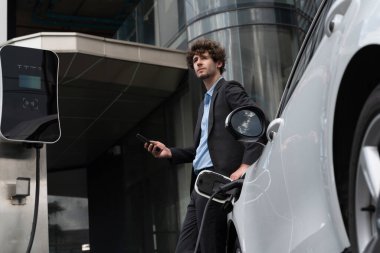 Progressive businessman talking on the phone, leaning on electric car recharging with public EV charging station, apartment condo residential building on the background as green city lifestyle.