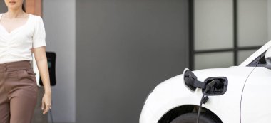 Focus image of electric vehicle recharging battery at home charging station with blurred woman walking in the background. Progressive concept of green energy technology applied in daily lifestyle.