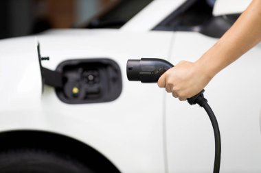 Focus hand holding EV charger plug with blurred background of progressive electric vehicle and socket parking in home garage with electric charging station powered by clean and sustainable energy.