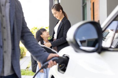 Closeup focus hand insert EV charger plug to electric vehicle at home charging station with blurred family in background. Progressive concept of green and clean energy to reduce CO2 emission by EV car