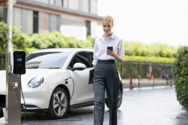 İş kadını tablet kullanıyor, halka açık otoparkta elektrikli aracını şarj ederken yürüyor. Teknolojinin ilerici yaşam tarzı ve EV otomobilinin ekolojik kaygısı