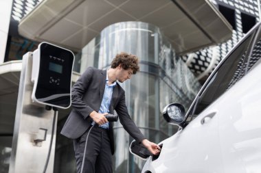 Below view closeup image of progressive black suit businessman recharge battery of his electric vehicle from public charging station. Renewable and alternative energy powered car concept.