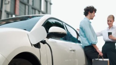 İlerici iş adamı ve iş kadını şehir merkezinde dolaşmadan önce şarj istasyonuna bağlı elektrikli arabaya yaslanıyor. Çevre dostu şarj edilebilir araba temiz enerjiyle çalışıyor..