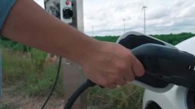 Elektrikli arabasıyla ilerici bir adam, EV arabası gelecekteki sürdürülebilir enerji kavramı olarak rüzgar türbiniyle yeşil alandaki enerji şarj etme istasyonundan enerji yüklüyor. Enerji üreteci olan elektrikli bir araç..