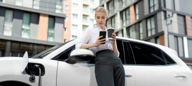 Focus businessman using phone, leaning on electric vehicle, holding coffee with blurred city residential condo buildings in background as progressive lifestyle by renewable and sustainable EV car.
