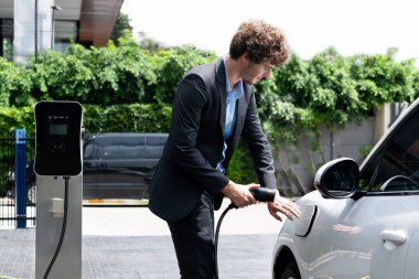Modern şehir yerleşim bölgesindeki kamu şarj istasyonunda elektrikli araba şarj eden yenilikçi bir işadamı. Çevre dostu şarj edilebilir araba alternatif temiz enerjiyle çalışıyor..