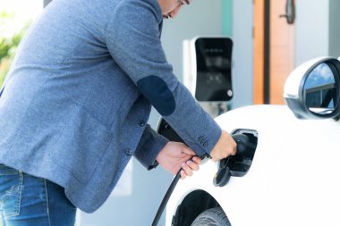 Progressive asian man install cable plug to his electric car with home charging station in the backyard. Concept use of electric vehicles in a progressive lifestyle contributes to clean environment.