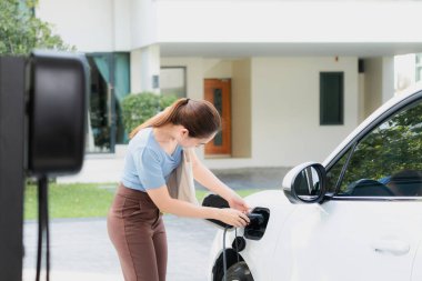 Progressive woman install cable plug to her electric car with home charging station. Concept of the use of electric vehicles in a progressive lifestyle contributes to clean environment.