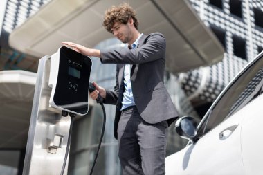 Below view of progressive businessman with electric car recharging at public charging station at modern city residential building. Eco friendly rechargeable EV car powered by alternative clean energy.