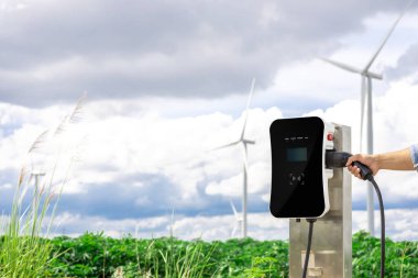 Progressive natural scenic with windmill generator where hand insert charging plug to electric vehicle from charging station with natural background. EV car powered by wind turbine electric generator.