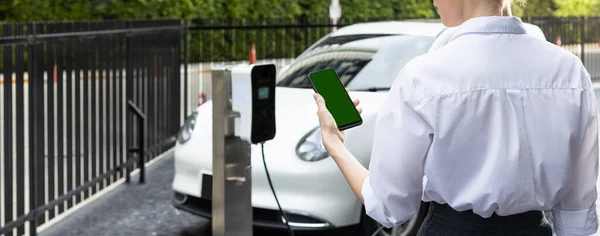 充電ステーションに電源ケーブルプラグと再生可能エネルギー駆動の電気自動車を設置しながら Ev車のバッテリー状態をモックアップ電話から見ているスーツクラッドの進歩的なビジネス女性 — ストック写真