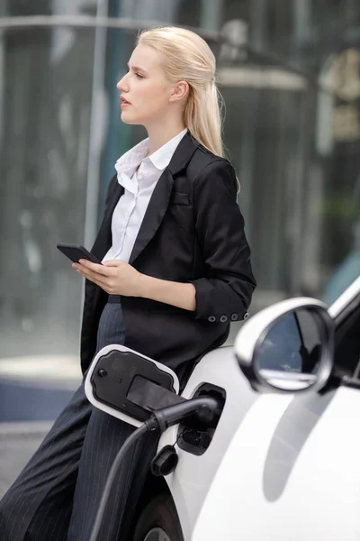 Businesswoman Wearing Black Suit Using Smartphone Leaning Electric Car Recharge — Stock fotografie