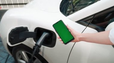 Taklit uzay için şarj istasyonuna bağlı elektrikli aracın yeşil ekran enerji durumunu gösteren bir model telefon. Temiz çevre için ilerici bir kavram. EV, yeşil yenilenebilir enerjiden güç alıyor.