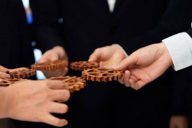 Closeup hand holding wooden gear by businesspeople wearing suit for harmony synergy in office workplace concept. Group of people hand making chain of gears into collective form for unity symbol. clipart