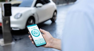 Closeup progressive businessman look at EV cars battery status application on smart phone screen at public parking charging station with power cable plug and renewable energy-powered electric vehicle