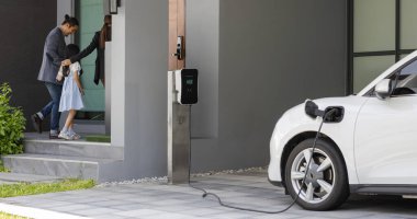 Progressive young parents and daughter with electric vehicle and home charging station. Green and clean energy from electric vehicles for healthy environment. Eco power from renewable source at home.
