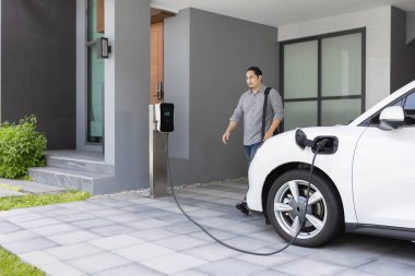 Progressive asian man and electric car with home charging station. Concept of the use of electric vehicles in a progressive lifestyle contributes to a clean and healthy environment.