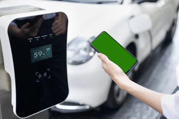 コピースペースの充電ステーションに接続された電気自動車の緑の画面表示エネルギーステータスを持つモックアップ電話 きれいな環境のための進歩的な概念 グリーン再生可能エネルギーを搭載したEv — ストック写真