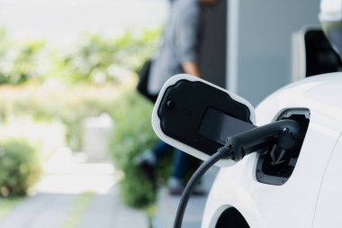 Focus electric car charging at home charging station with blurred progressive man walking in the background. Electric car using renewable clean for eco-friendly concept.