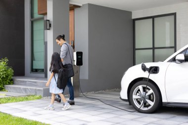 Progressive father and daughter plugs EV charger from home charging station to electric vehicle. Future eco-friendly car with EV cars powered by renewable source of clean energy.