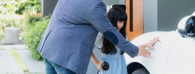 Progressive father and daughter returned from school in electric vehicle that is being charged at home. Electric vehicle driven by renewable clean energy. Home charging station concept for environment