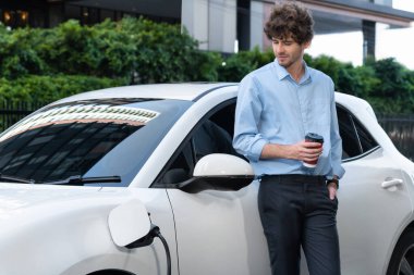 Progressive eco-friendly concept of parking EV car at public electric-powered charging station in city with blur background of businessman leaning on recharging-electric vehicle with coffee.