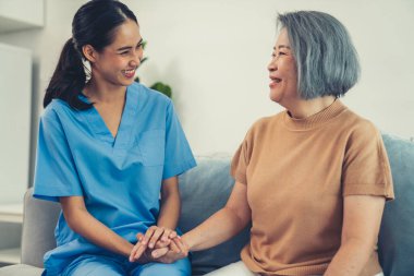A caregiver rest her hands on the shoulders of a contented senior patient while she sitting on the sofa at home.