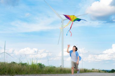 İlerici mutlu kaygısız çocuk konsepti. Yol boyunca koşan ve uçurtma uçuran genç bir çocuk kırsal alanda doğal manzaralı, açık gökyüzü ve güneşli bir gün dağ ve rüzgar türbini arka planında