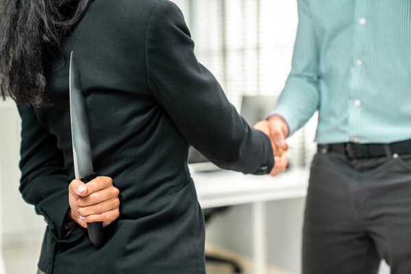 Back view of businesswoman shaking hands with another businessman while holding a knife behind his back. Concept of back backstabbing in business, backstabbing between colleagues.