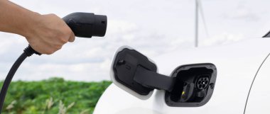 Progressive natural scenic with windmill generator where hand insert charging plug to electric vehicle from charging station with natural background. EV car powered by wind turbine electric generator.
