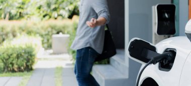 Focus electric car charging at home charging station with blurred progressive man walking in the background. Electric car using renewable clean for eco-friendly concept.