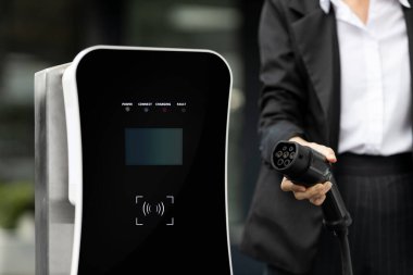 Focus EV charger plug and electric car at public charging station with blur progressive businesswoman holding charger and apartment condo building in background. Eco friendly electric vehicle concept.