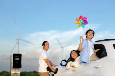 İlerici mutlu aile kavramı elektrikli araçla rüzgar çiftliğinde eğleniyor. Rüzgar türbini jeneratöründen gelen temiz yenilenebilir enerji ile şarj edilen elektrikli araç..