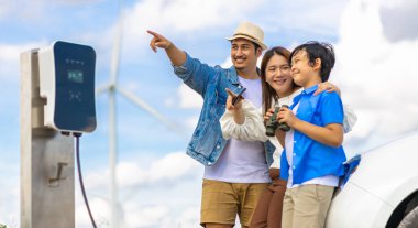 İlerici mutlu aile kavramı elektrikli araçla rüzgar çiftliğinde eğleniyor. Rüzgar türbini jeneratöründen gelen temiz yenilenebilir enerji ile şarj edilen elektrikli araç..