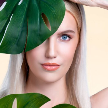 Closeup facial portrait personable woman with perfect smooth makeup holding green monstera leaves and cover her face as natural healthy skincare treatment. Tropical nature and beauty concept