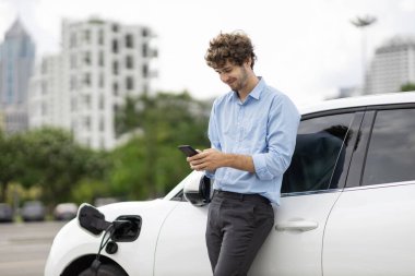 Progressive businessman talking on the phone, leaning on electric car recharging with public EV charging station, apartment condo residential building on the background as green city lifestyle.