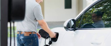 Progressive asian man install cable plug to his electric car with home charging station in the backyard. Concept use of electric vehicles in a progressive lifestyle contributes to clean environment.