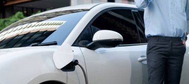 Closeup progressive suit-clad businessman with his electric vehicle recharge his car on public charging station in modern city with power cable plug and renewable energy-powered electric vehicle.