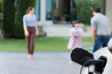 Focus closeup electric vehicle recharging battery from home electric charging station with blurred family in background. Renewable clean energy car for progressive eco awareness lifestyle concept.