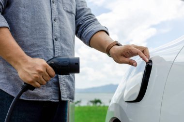 Progressive natural scenic where hand insert charging plug to electric vehicle from charging station with natural background. Future energy concept of EV car powered by sustainable electric energy.