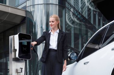 Progressive businesswoman wearing suit with electric car recharging at public parking car charging station at modern city center. Eco friendly rechargeable car powered by alternative clean energy.