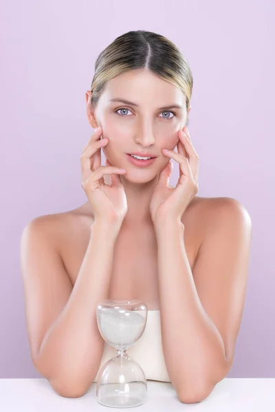 Alluring beauty model using hourglass in beauty concept of anti-aging skincare treatment for woman. Beautiful caucasian women portrait with perfect smooth clean skin in pink isolated background.