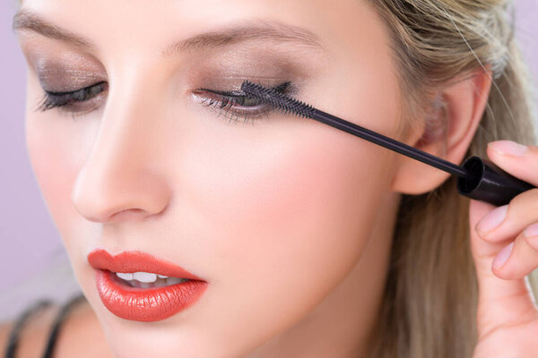 Женщина крупным планом с светлыми волосами кладет заманчивую черную тушь с кистью в руке на длинные густые ресницы. Идеально модная косметическая чистая кожа лица с красивым глазом молодая женщина в высоком разрешении.